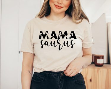 A woman stands indoors, smiling with one hand on her waist, wearing an InkMerge Mama Saurus t-shirt featuring Mama Saurus in bold and script fonts.